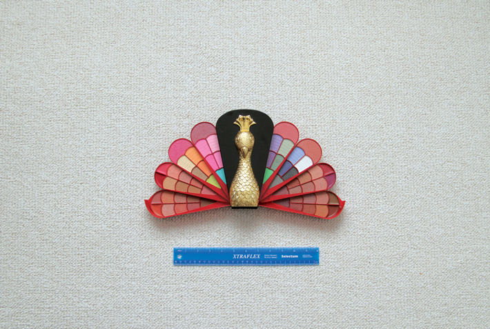 A photograph of a makeup palette in the shape of a peacock being measured by a ruler.