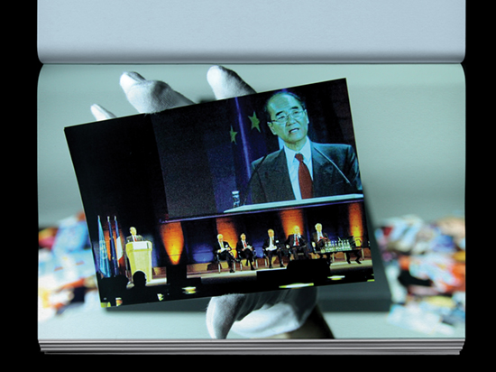 A hand wearing a white archive glove holds a photograph of people sitting on a stage with a large portrait behind them.