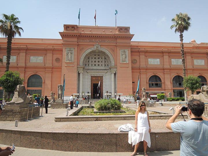 A man taking a photograph of a woman in front of a large pink building with palm trees and little sphinx of Giza sculptures on either side of it.