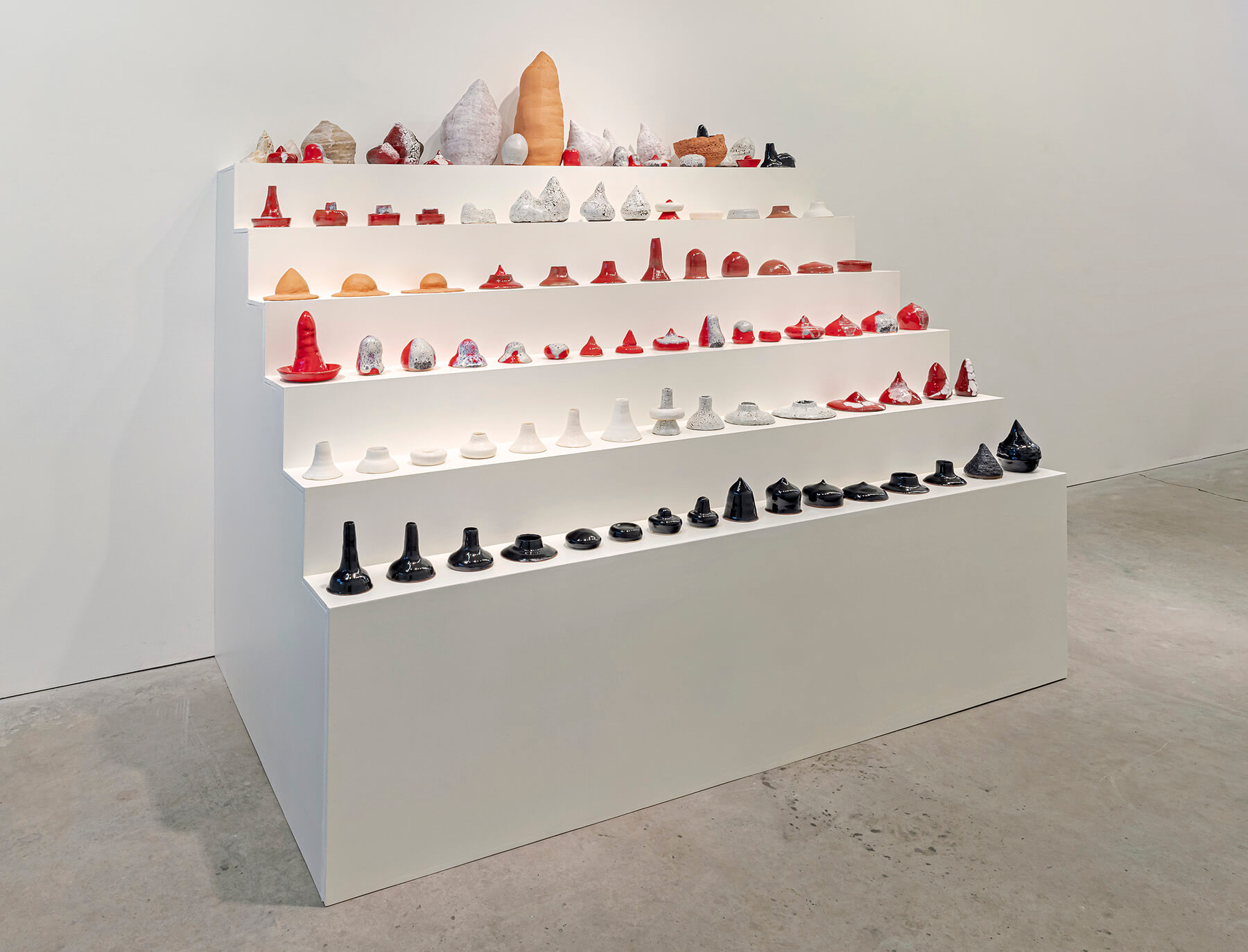 Full installation view of all sculptures lined up on a six level white plinth shaped like stair steps (side).