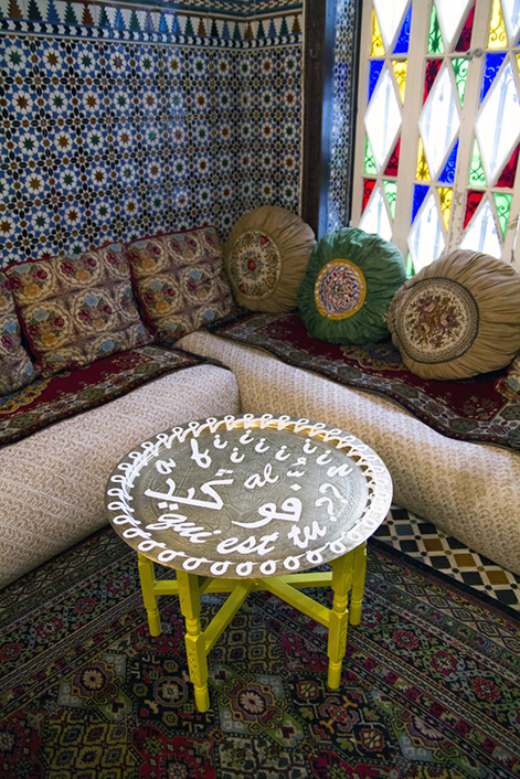 A round coffee table standing on an oriental rug, next to a patterned couch with various pillows against a wall with a repeating pattern and a stain glass window.