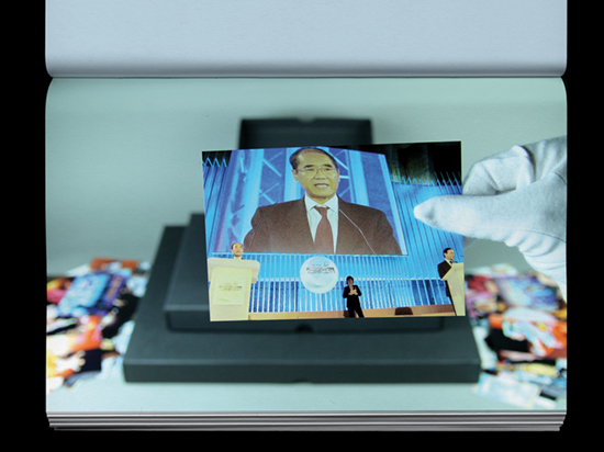 A hand wearing a white archive glove holds a photograph of three people standing on a stage with a large portrait behind them.