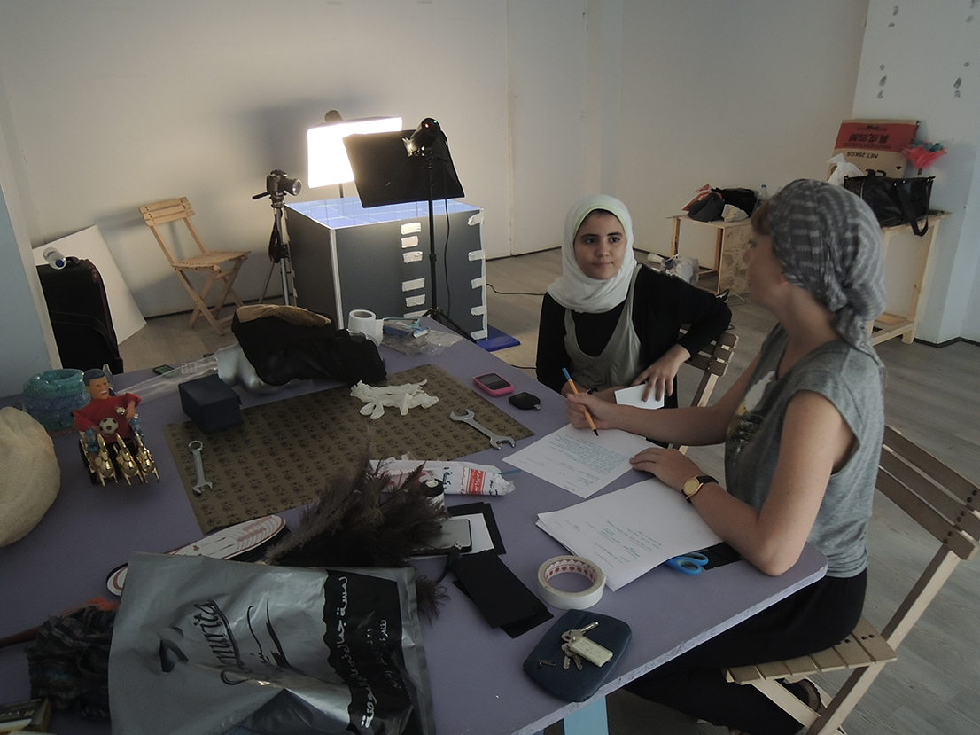 Two people sit at a table writing notes, surrounded by objects.