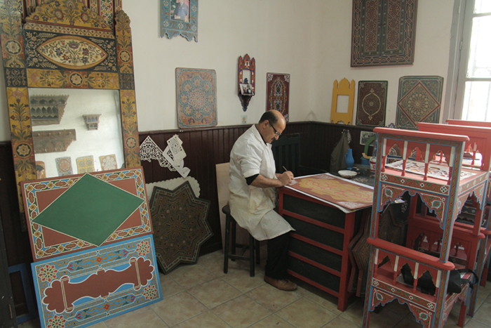 A man sits in a room surrounded by art pieces, while drawing on a new art piece.