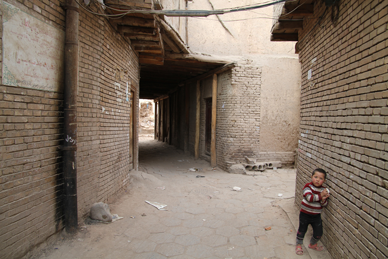A photograph on ground level between two brick buildings with a child leaning against one of them staring into the lens of the camera.