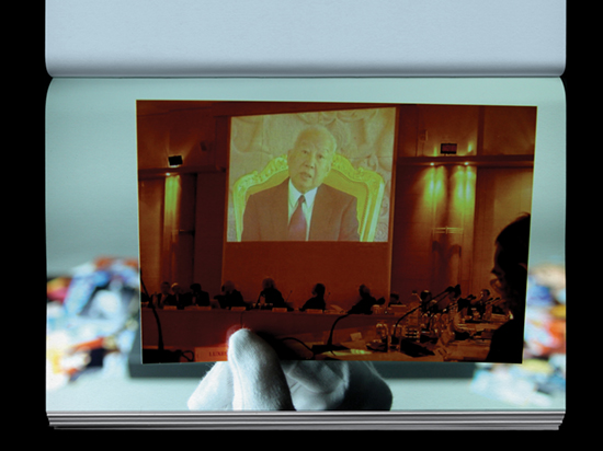 A hand wearing a white archive glove holds a photograph of four people sitting on a stage with a projection of photograph of a man behind them.
