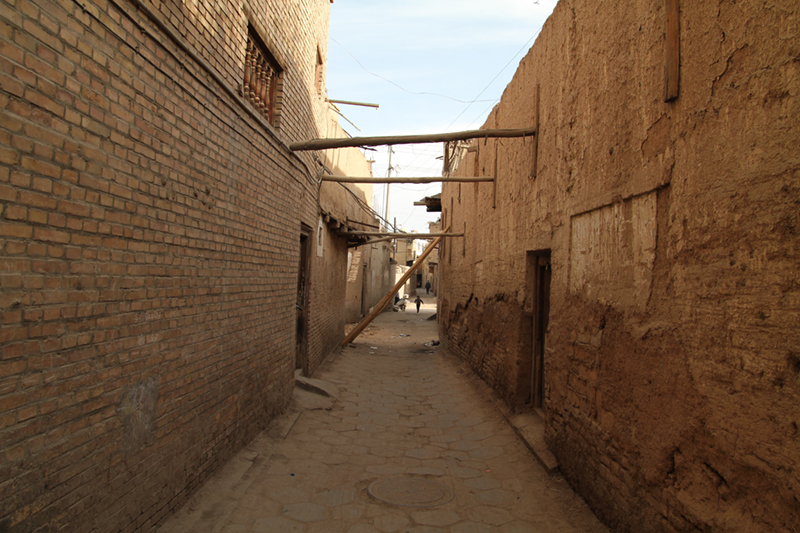 A photograph on the street between two buildings with wooden beams above, one of them has fallen.