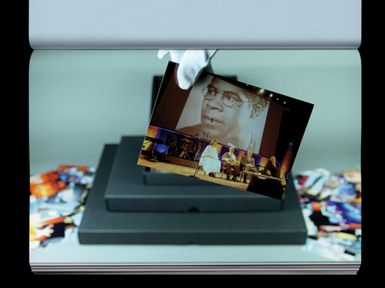 A hand wearing a white archive glove holds a photograph of four people sitting on a stage with a projection of photograph of a man behind them.