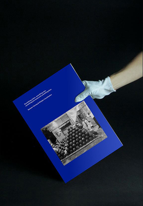 A hand wearing a white archiving glove holds the blue publication against a black background.