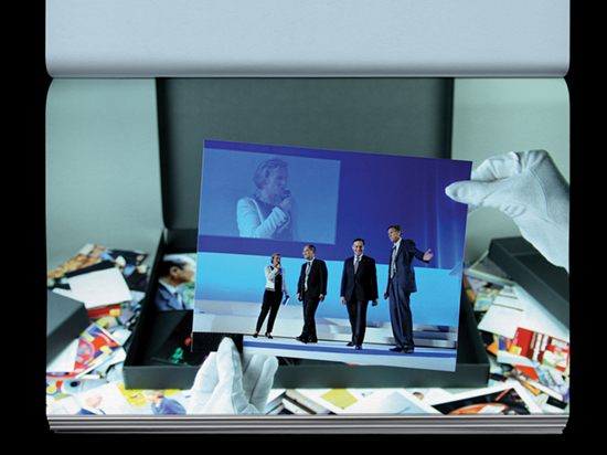 Hands wearing white archive gloves holding a photograph of four people standing on a stage with a projection of one of the people behind them.