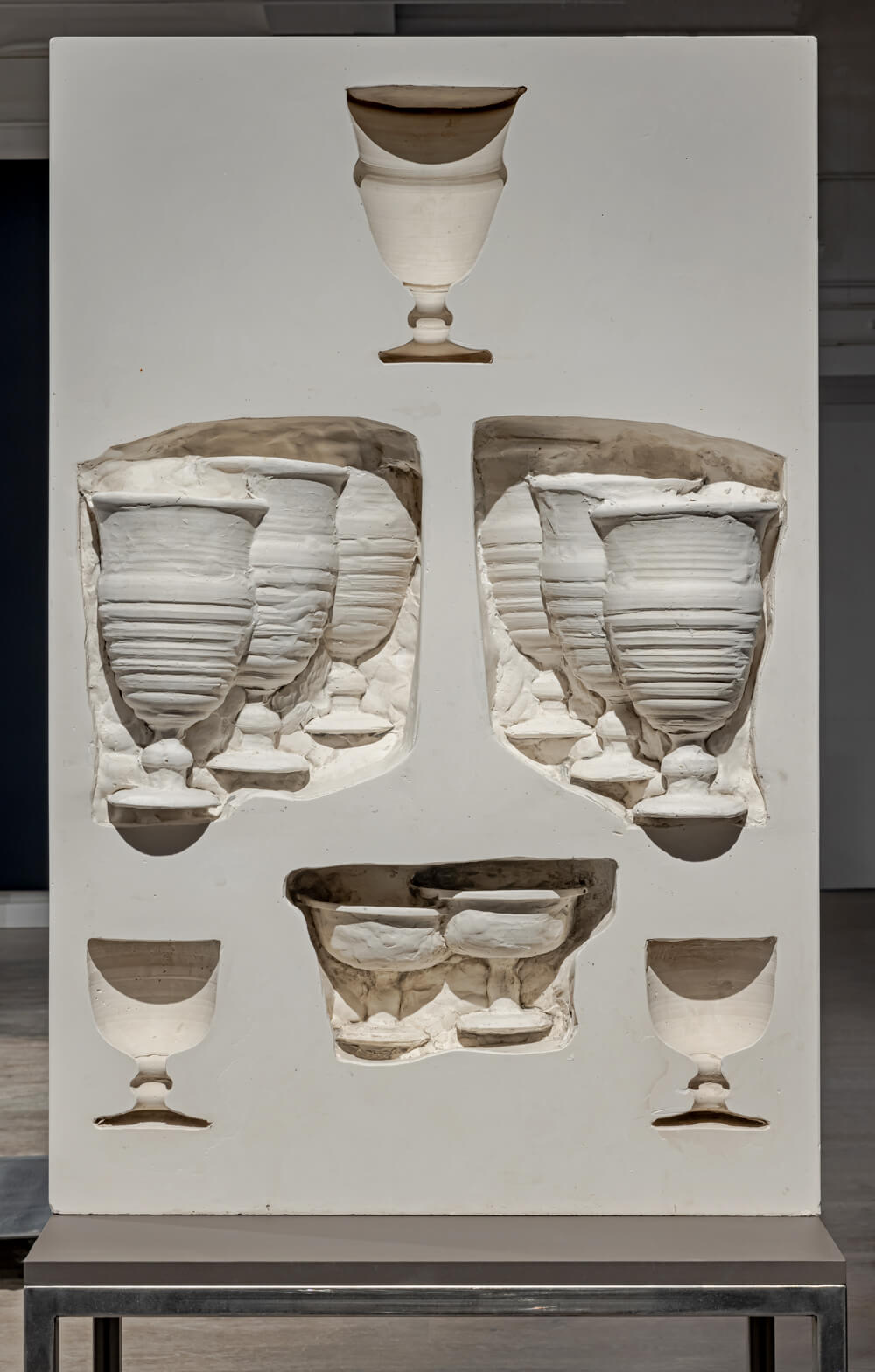 Clay wall with recessed areas shaped like Goblets. Some of the goblets are inside the recessed areas.
