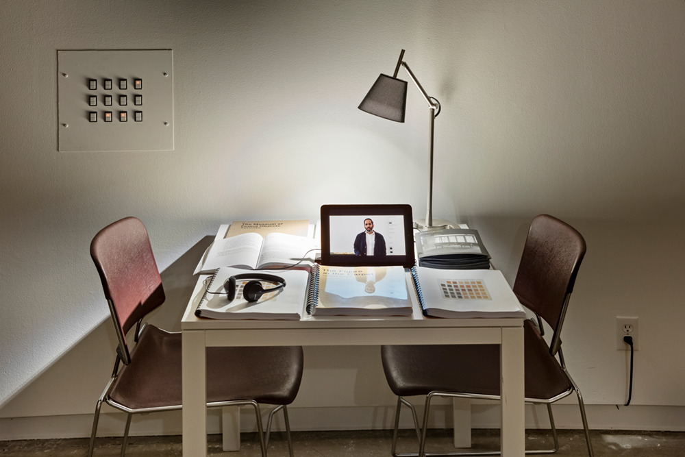 Open books, notebooks, and a propped up tablet with headphones sit on top of a white desk with two chairs on either side of it.