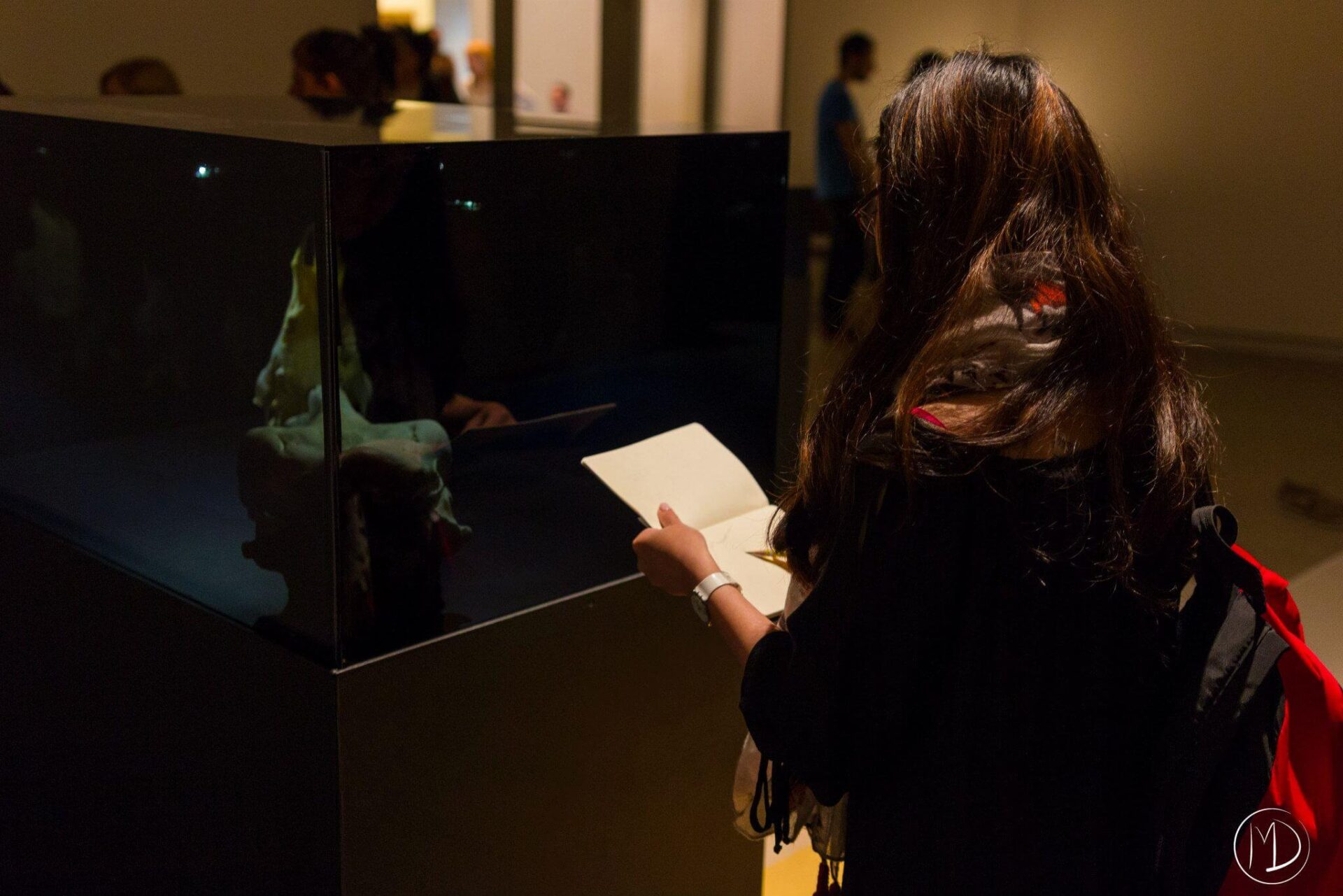 A person sketching in their note book while looking at a piece inside of a glass case.