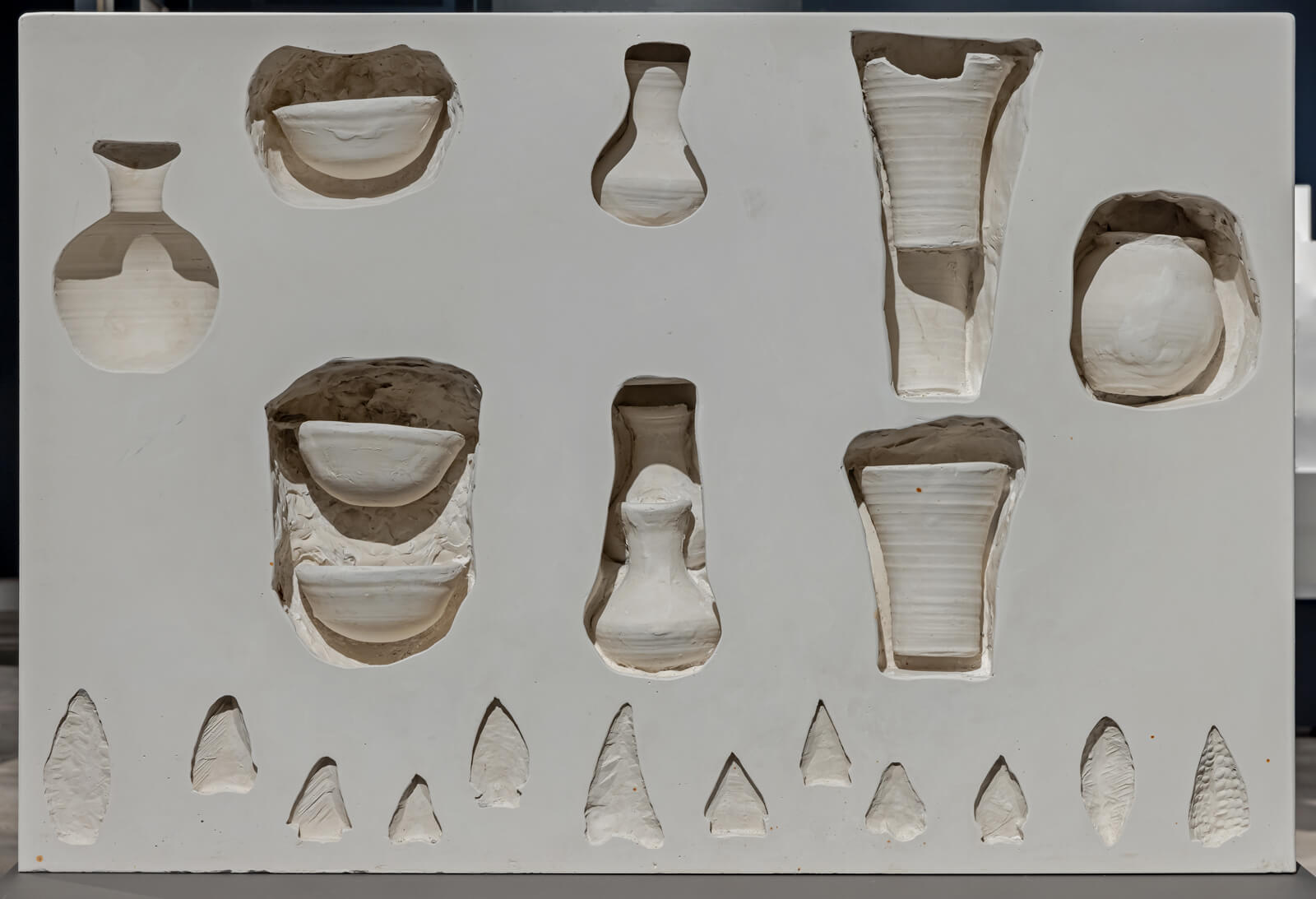 Clay wall with recessed areas shaped like bowls, vases, and spears. Most of the pottery is inside the recessed areas.