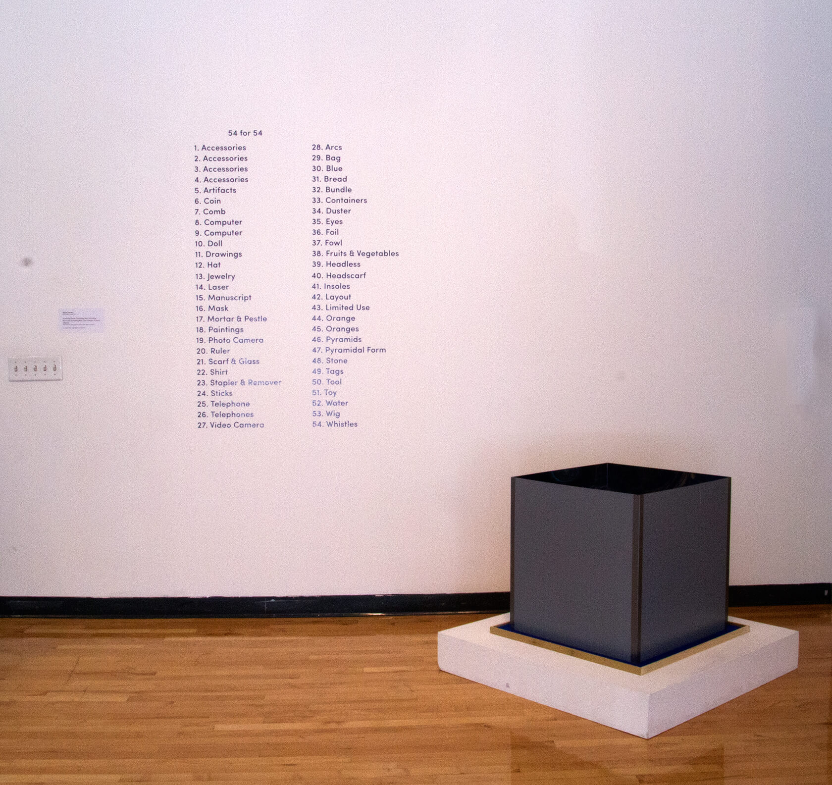 A list of items set in black vinyl text on a white gallery wall, a dark grey box on a plinth stands next to it.