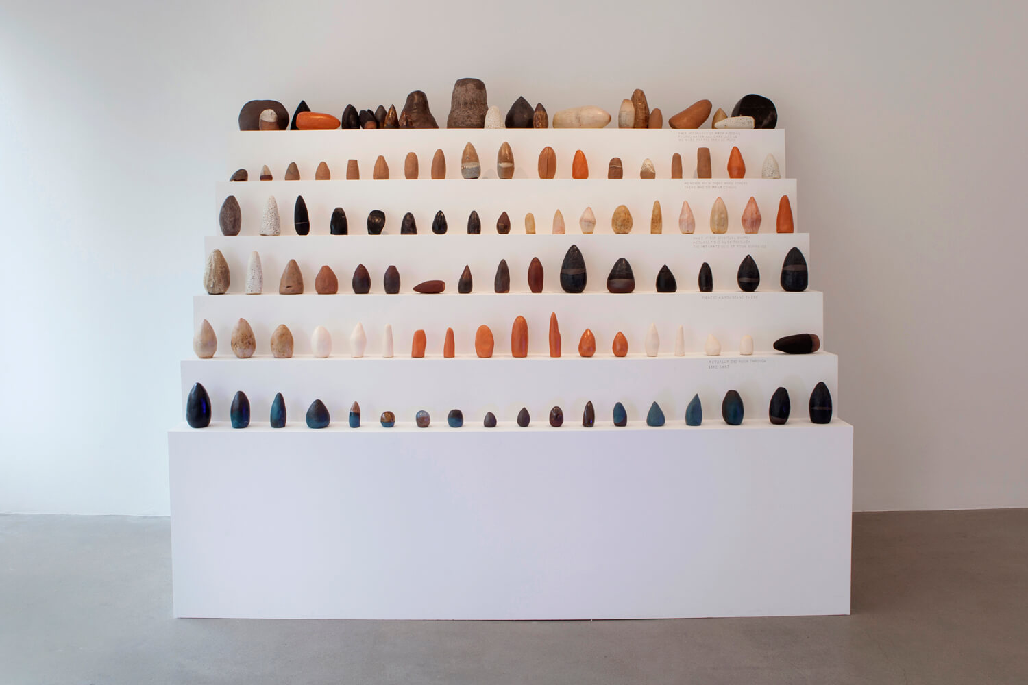 Full installation view of all sculptures lined up on a six level white plinth shaped like stair steps (front).