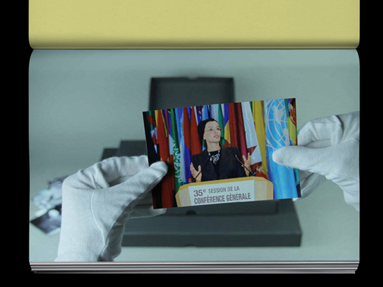 A hand wearing a white archive glove holds a photograph of a person making hand gestures as they speak into a microphone behind a podium.