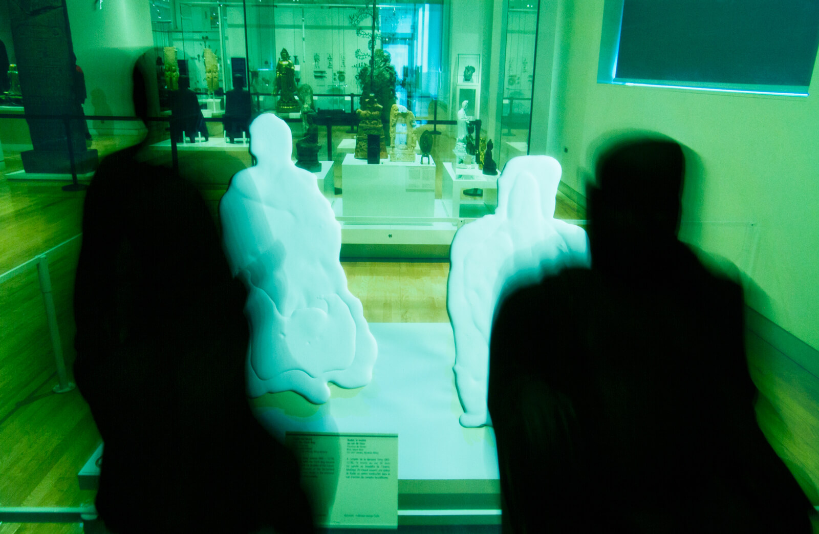 Two body shaped sculptures in a museum setting with a silhouette of two human figures in motion in the foreground.