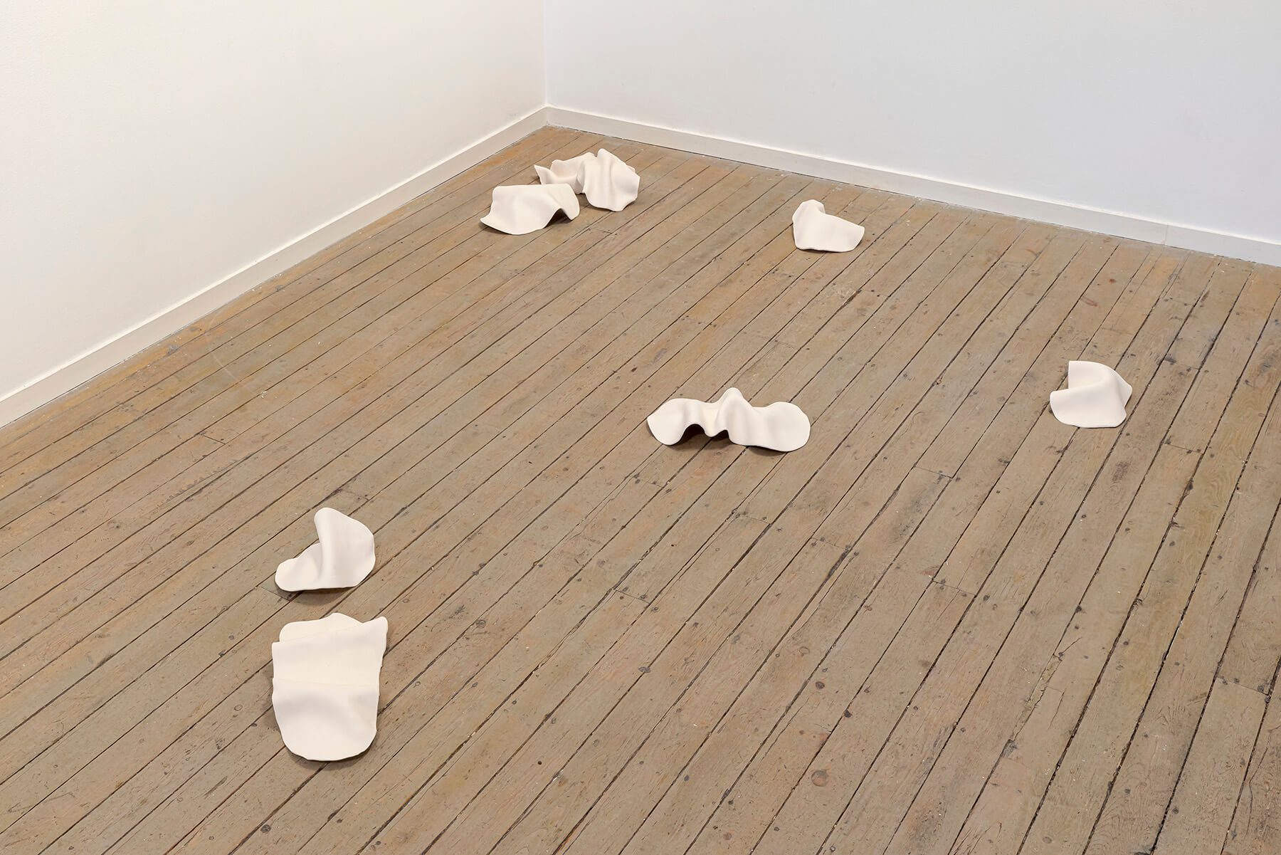 Dispersed clay sculptures shaped like veils lying on a wooden floor.
