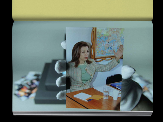 A hand wearing a white archive glove holds a photograph of a woman sitting behind a desk making a gesture with her hands.
