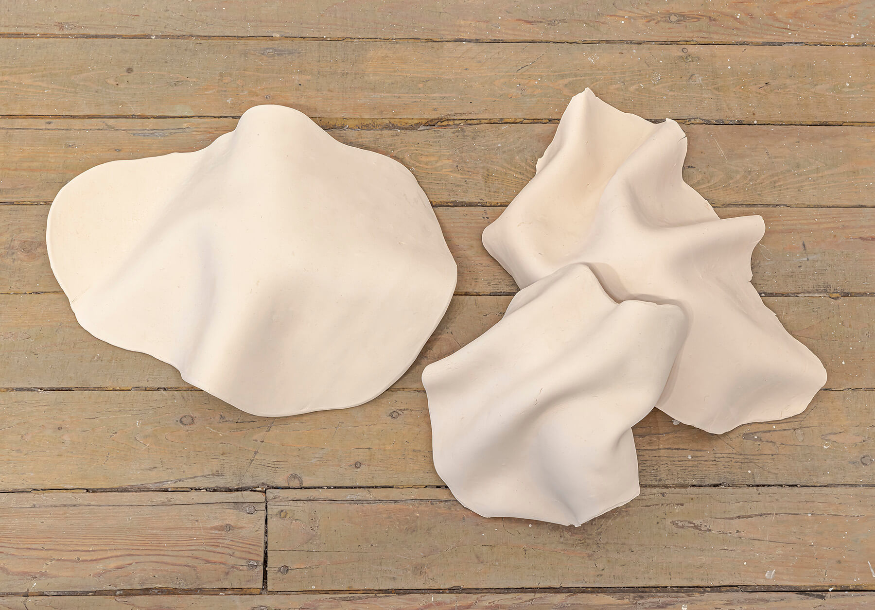 Three clay sculptures shaped like veils lying on a wooden floor.