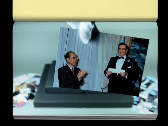 A hand wearing a white archive glove holds a photograph of a short and tall person wearing suits in a formal setting.