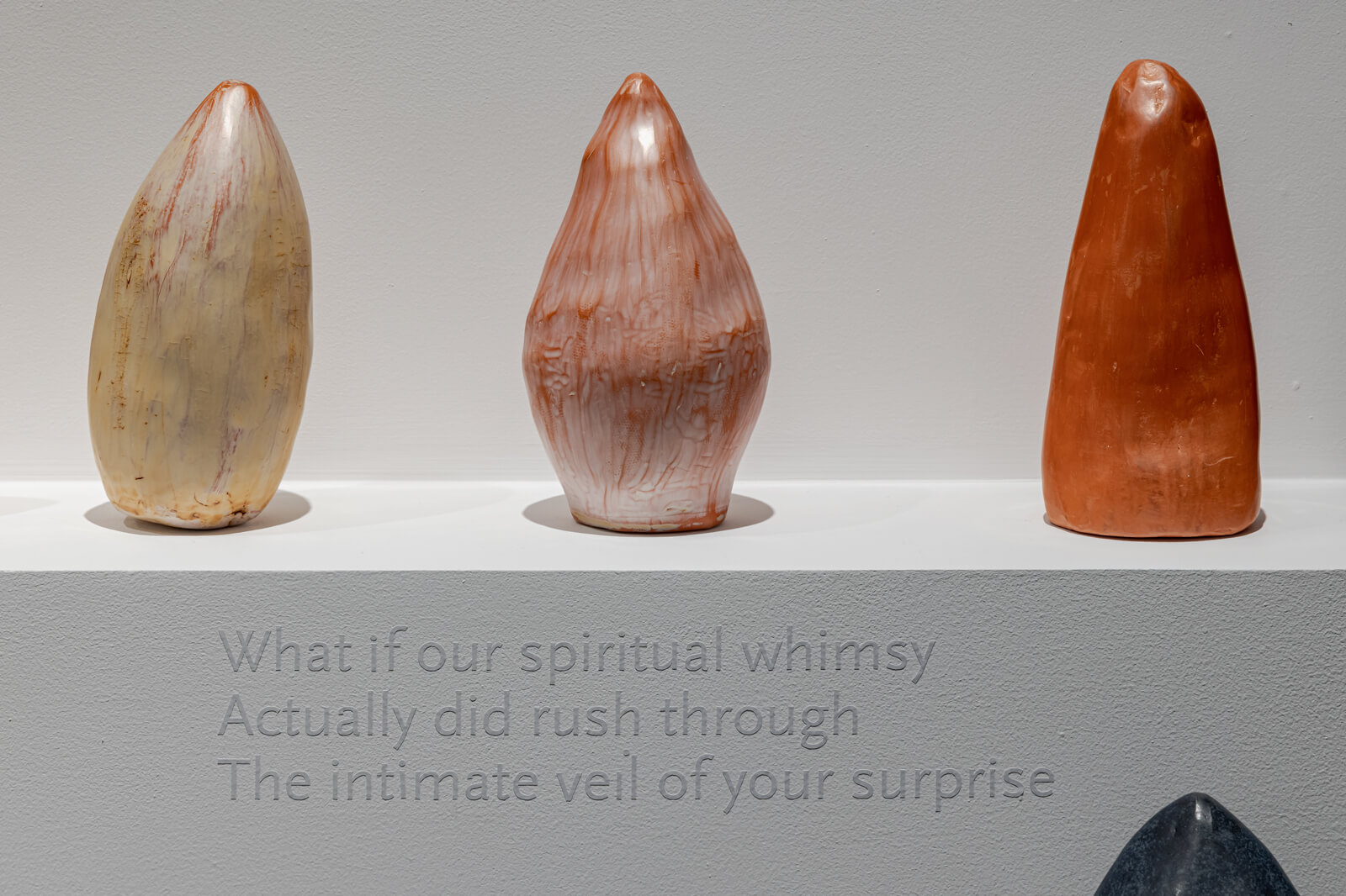 Three oblong sculptures of similar size lined up on a white plinth, with vinyl text underneath it.