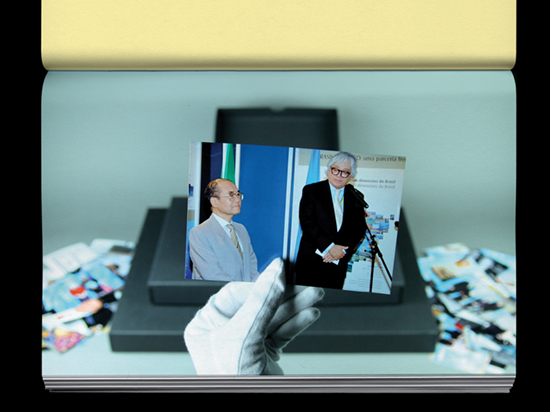 A hand wearing a white archive glove holds a photograph of a tall and short person in suits and ties standing in front of a microphone.