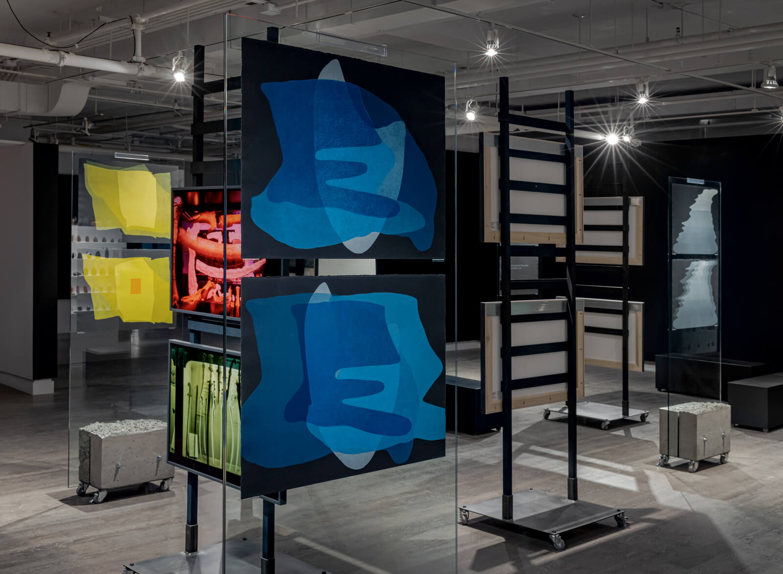 Installation view of blue prints hanging on a glass panel, with other prints in the background hung in a similar manner.