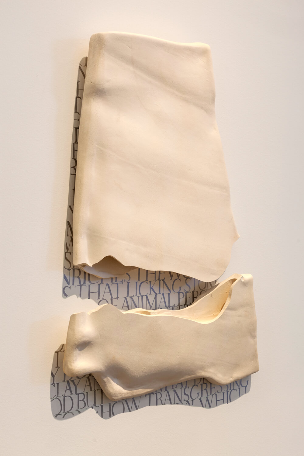 Two pieces of a white clay tablet hanging on a white wall, they cast shadows on top of black text written on the wall to fit the exact area of the shadow.