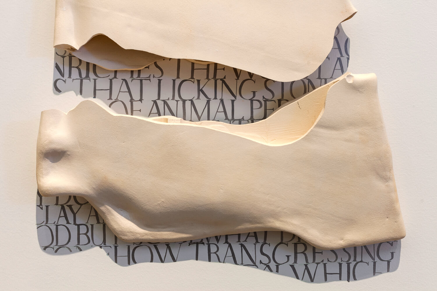 Two pieces of a white clay tablet hanging on a white wall, they cast shadows on top of black text written on the wall to fit the exact area of the shadow.