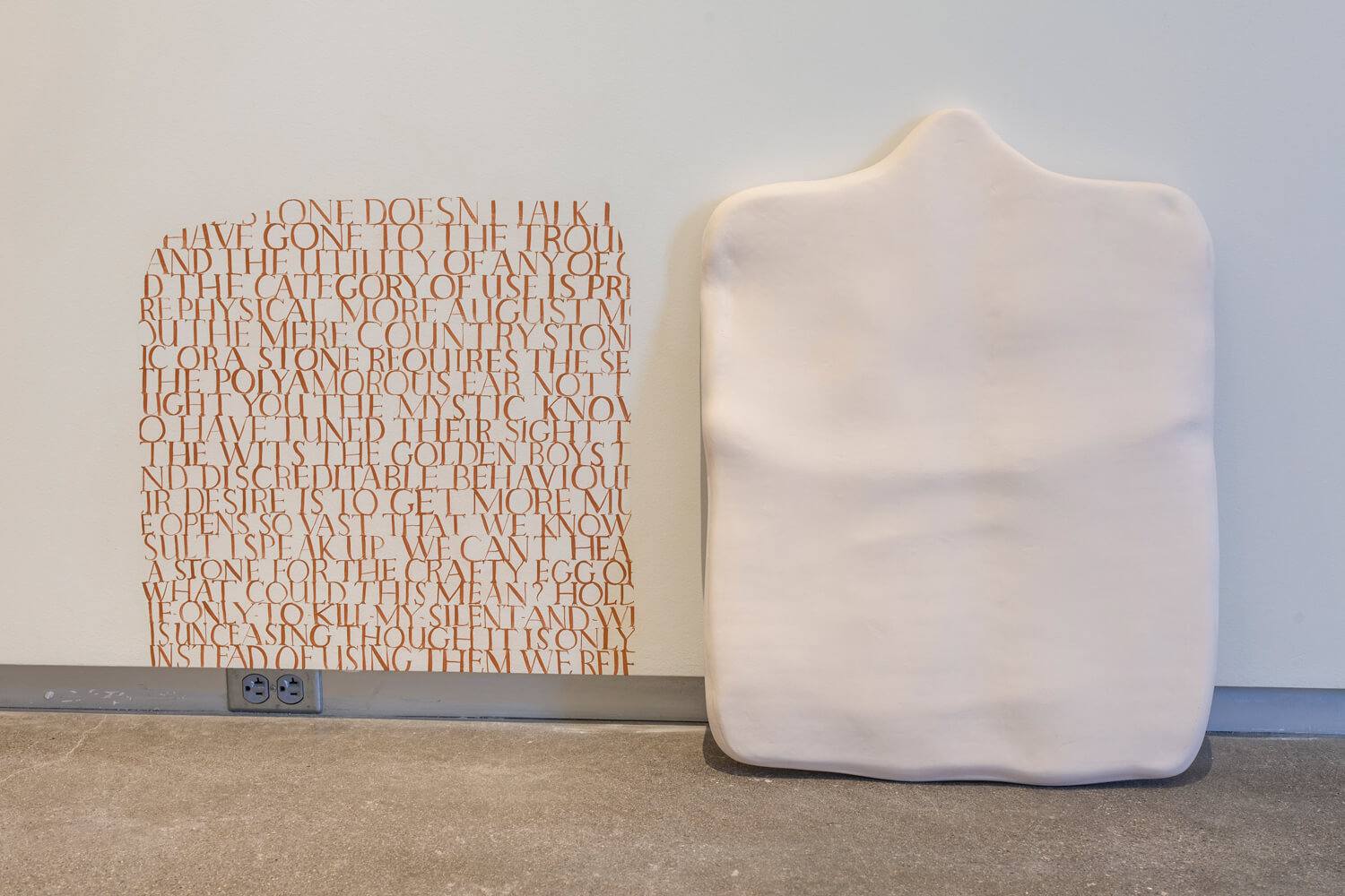 A white clay tablet leaning against a white wall next to a paragraph of the same shape and size on the wall, containing hand written capitalized dark orange text with no leading.